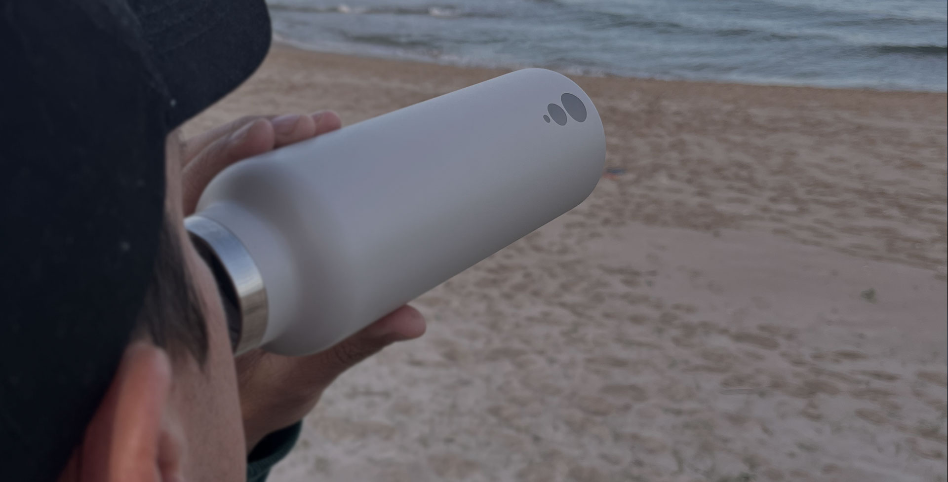 Una persona sostiene una botella WaterGo blanca frente al mar. Sobre el acero se ven grabados los tres círculos del logotipo.