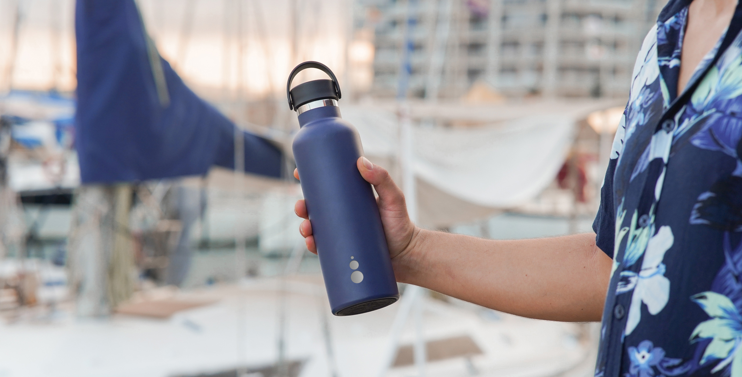 Una mano sostiene una botella WaterGo azul en un puerto deportivo; en el acero se ven grabados los tres círculos del logotipo.