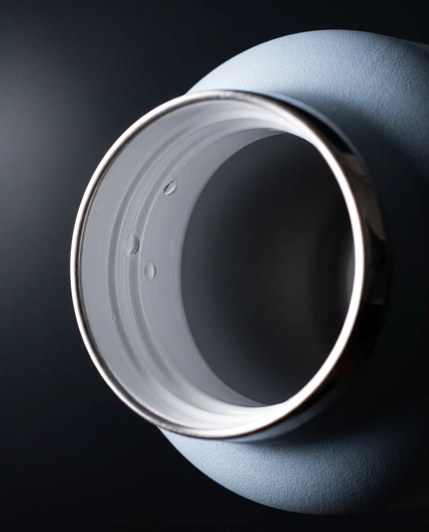 Macro de la boca de una botella WaterGo vista de lado: el collarín de acero pulido rodea un interior de cerámica blanca y brillante, con gotas de agua adheridas.