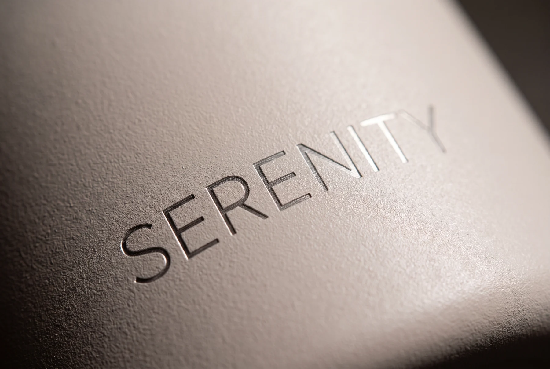 Macro del grabado láser de la palabra SERENITY sobre el acabado nude: el láser ha quitado la pintura y el trazo brilla en el acero desnudo, con la textura mate alrededor.